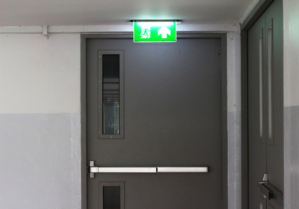 A grey fire door with a push bar and illuminated green emergency exit sign above it in a commercial corridor.